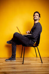 Gut aussehender Mann, der mit einem Laptop auf einem Stuhl sitzt und wegschaut - DAWF01617