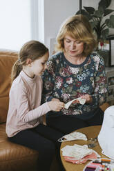 Mature woman with granddaughter making protective face mask at home during pandemic - VBF00099