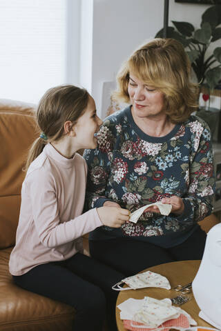 Smiling girl looking at grandmother holding homemade face mask in living room stock photo