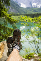 Italy, Province of Udine, Tarvisio, Feet of man wearing hiking boots relaxing on shore of Fusine Lake - GIOF08413