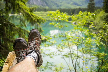 Italy, Province of Udine, Tarvisio, Feet of man wearing hiking boots relaxing on shore of Fusine Lake - GIOF08414