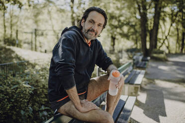 Selbstbewusster reifer Mann, der eine Wasserflasche hält, während er auf einer Bank im Park sitzt - JLOF00451