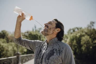 Nahaufnahme eines reifen Mannes, der Wasser trinkt, während er in einem Park vor einem klaren Himmel steht - JLOF00479