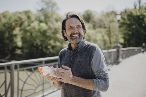 Smiling man holding water bottle while standing on footbridge in park - JLOF00484