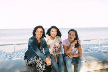 Smiling mother with daughters sitting on retaining wall against sea - DCRF00300
