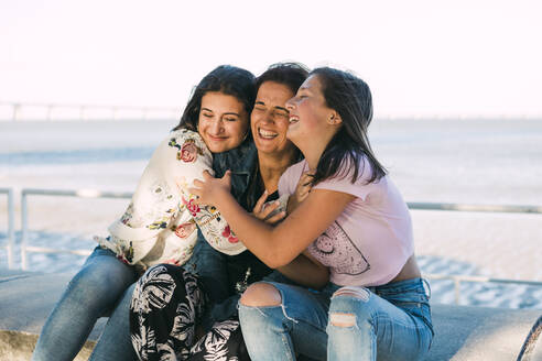 Liebevolle Töchter, die ihre fröhliche Mutter umarmen, während sie auf einer Stützmauer am Meer sitzen - DCRF00301