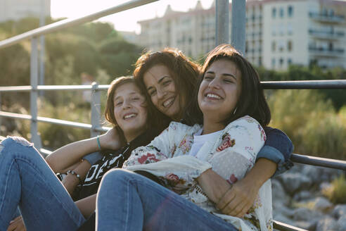 Lächelnde Mutter mit Tochter am Geländer sitzend bei Sonnenuntergang - DCRF00316