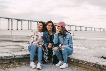 Smiling mother with daughters sitting on retaining wall against sea - DCRF00319