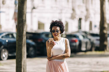 Stilvolle Frau mit lockigem Haar, die ihr Smartphone benutzt, während sie in der Stadt steht - DCRF00322