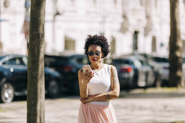 Stylish woman with curly hair using smart phone while standing in city - DCRF00322