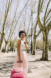 Smiling woman holding sunglasses with suitcase standing on footpath in city - DCRF00326