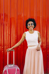 Smiling woman wearing sunglasses with suitcase standing against orange wall - DCRF00330