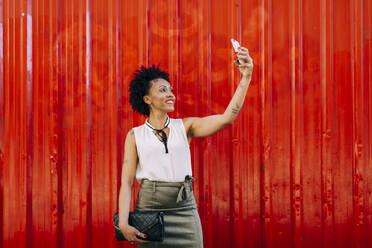 Lächelnde mittelgroße erwachsene Frau, die ein Selfie mit ihrem Smartphone macht, während sie an einer orangefarbenen Wand steht - DCRF00333
