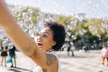 Fröhliche erwachsene Frau spielt mit Seifenblasen in der Stadt - DCRF00339