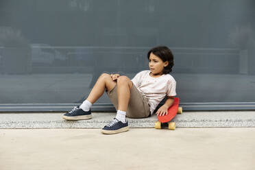 Thoughtful boy with skateboard relaxing on footpath against wall - VABF03066