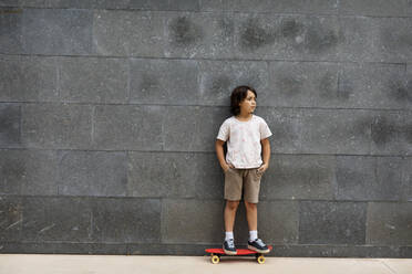 Nachdenklicher Junge mit Händen in den Taschen, der auf einem Skateboard an der Wand steht - VABF03067