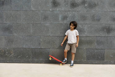 Boy playing with skateboard while standing on footpath against wall - VABF03068
