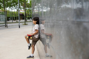 Boy holding skateboard looking away while standing by wall in park - VABF03069