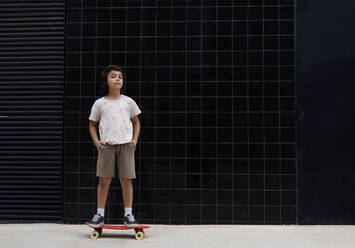 Junge mit Händen in den Taschen steht auf Skateboard gegen die Wand - VABF03070