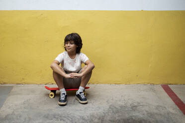 Thoughtful boy sitting on skateboard against wall - VABF03077