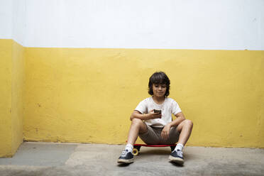 Boy using smart phone while sitting on skateboard against wall - VABF03078