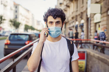 Portrait of young man wearing mask talking over smart phone while standing in city - MEUF00896