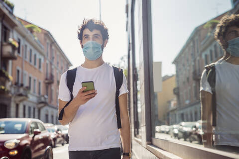 Man wearing mask holding smart phone while walking in city stock photo