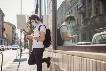 Young man wearing mask using smart phone while standing by built structure in city - MEUF00899