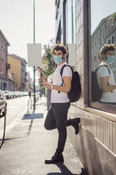 Man wearing mask using smart phone while standing by built structure in city - MEUF00900