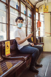 Junger Mann mit Maske, der in der Straßenbahn sitzt und sein Smartphone benutzt - MEUF00907