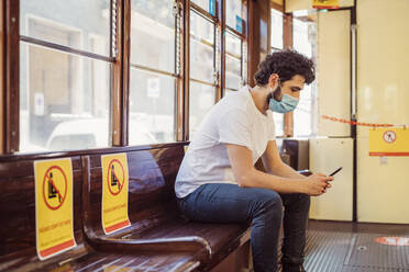 Mann mit Gesichtsmaske benutzt Mobiltelefon in der Straßenbahn - MEUF00909