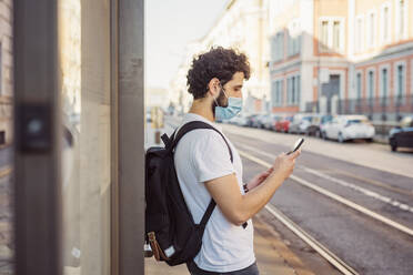 Mann mit Rucksack trägt Maske, während er in der Stadt ein Mobiltelefon benutzt - MEUF00916