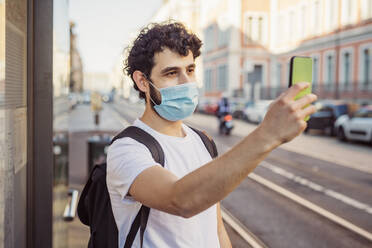 Junger Mann mit Maske, der ein Selfie mit seinem Smartphone macht, während er in der Stadt steht - MEUF00918