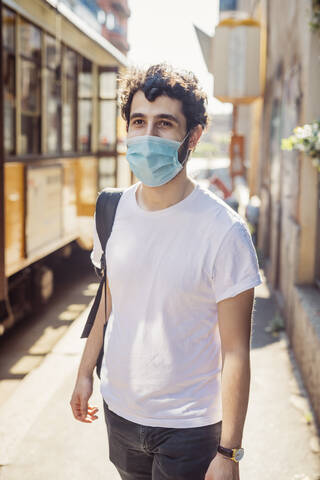 Junger Mann mit Maske, der an einem sonnigen Tag auf einer Straße in der Stadt spazieren geht, lizenzfreies Stockfoto