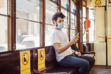 Junger Mann mit Gesichtsmaske, der in der Straßenbahn sitzt und sein Smartphone benutzt - MEUF00923