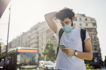 Junger Mann mit Gesichtsmaske, der sein Smartphone benutzt, während er an einem sonnigen Tag in der Stadt steht - MEUF00924