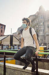 Junger Mann mit Maske schaut weg, während er auf einem Geländer in der Stadt sitzt - MEUF00926