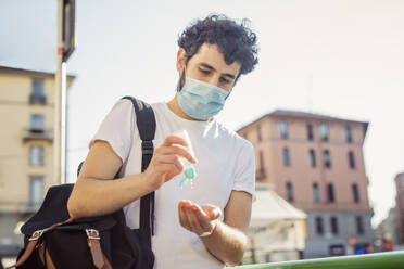 Man wearing face mask washing hands with sanitizer while standing against clear sky - MEUF00927