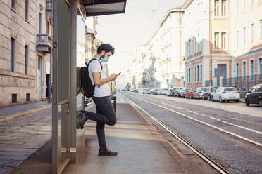 Mann mit Gesichtsmaske, der ein Smartphone benutzt, während er an einem Gebäude in der Stadt steht - MEUF00931