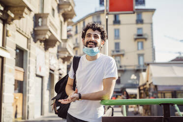 Smiling young man with face mask looking away while standing by railing in city - MEUF00935