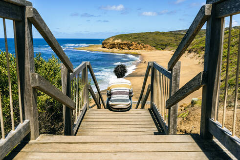 Mann schaut auf Bells Beach, während er auf einer Holztreppe sitzt, Victoria, Australien - KIJF03108