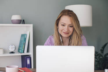Smiling businesswoman wearing headphones using laptop in office - MOMF00876