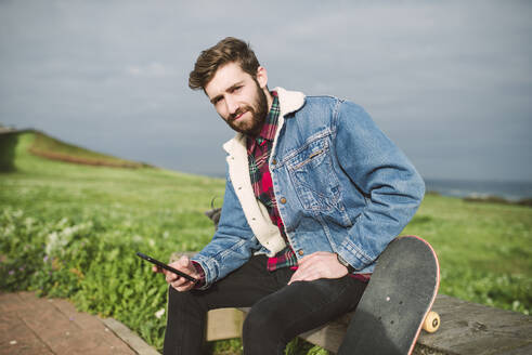 Gut aussehender Mann, der ein Smartphone benutzt, während er mit einem Skateboard auf dem Sitz gegen den bewölkten Himmel sitzt - RAEF02400