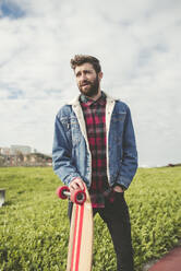 Bearded man holding skateboard while standing on land against cloudy sky in park - RAEF02406