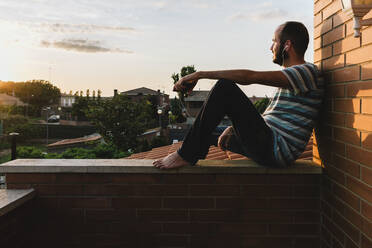 Nachdenklicher Mann, der Musik hört, während er auf einer Mauer sitzt und den Himmel bei Sonnenuntergang betrachtet - XLGF00210