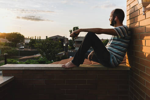 Thoughtful man listening music while sitting on retaining wall against sky during sunset - XLGF00210