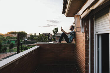 Mittelgroßer erwachsener Mann, der sein Smartphone benutzt, während er bei Sonnenuntergang auf der Terrasse an einer Stützmauer sitzt - XLGF00211