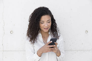 Geschäftsfrau mit lockigem Haar, die ein Smartphone benutzt, während sie an der Wand steht - JPTF00563