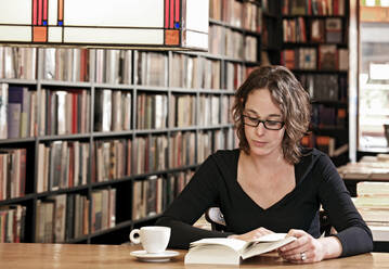 Woman drinking coffee while reading at bookstore - CAVF85508