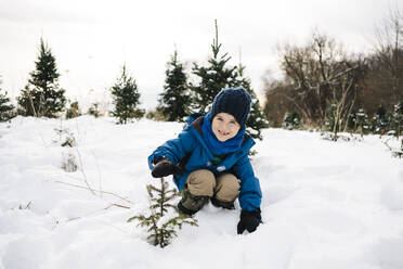 Lächelnder Junge zeigt Baum, während er auf schneebedecktem Land vor dem Himmel kauert - JVSF00013